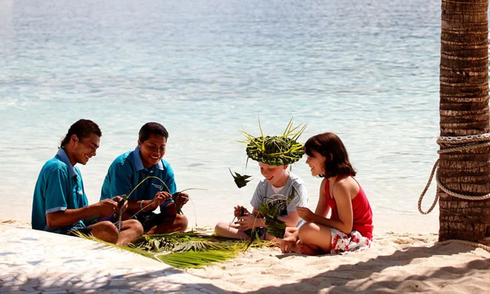 Two children sitting on the sand with two staff members learning the tradition of basket weaving at Plantation Island Resort
