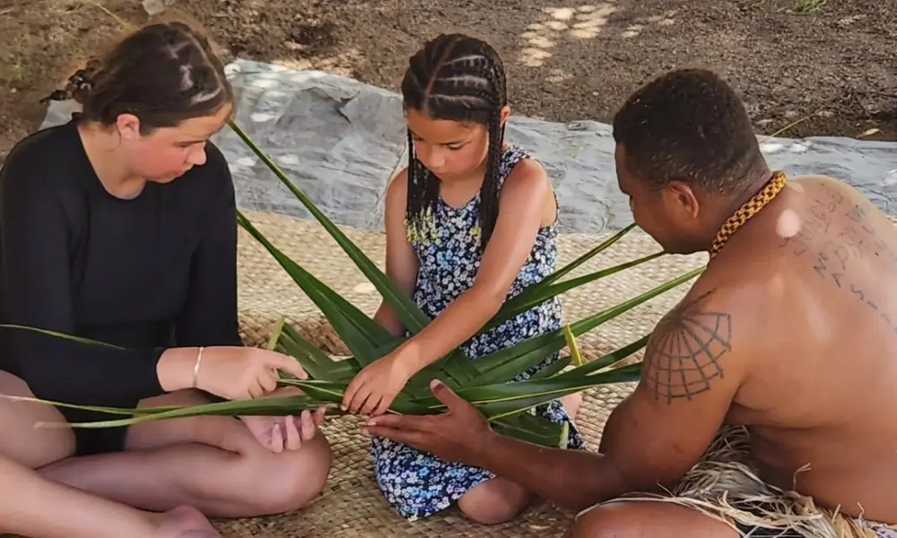 A child learns how to weave baskets with Fijian guide and parent
