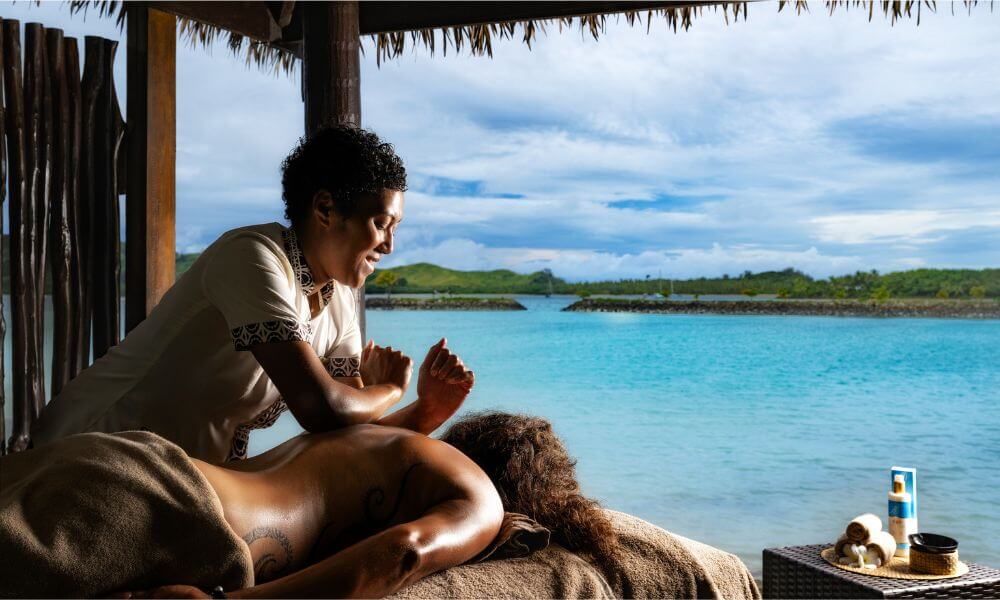 A spa therapist gives a back massage to a guest in an open-air hut overlooking a tropical turquoise lagoon.