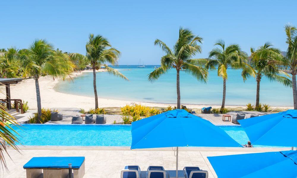 A bright blue resort swimming pool and sun umbrellas overlooking a white sand beach with palm trees and calm ocean water.