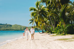 Bride & Groom walking along a white sand beach with palm trees and turquoise lagoon.